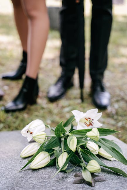 A delicate arrangement of white lilies with open petals displaying brown stamens and partially opened buds, complemented by elongated green leaves, rests on a grey stone surface. The bouquet appears f