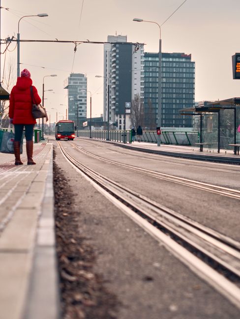 A person dressed in a bright red puffer jacket and dark trousers is standing on the tram stop platform near Addington Village tram stop, waiting as a tram approaches on the tracks. In the background, 