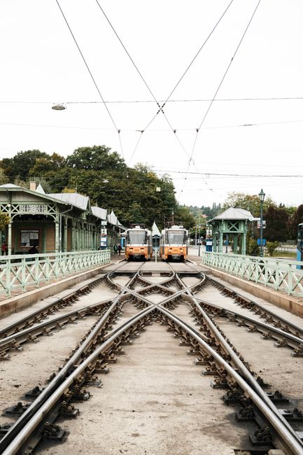 The image depicts a tram stop with two vintage-style trams facing each other on parallel tracks. The platform is flanked by light green wooden shelters with decorative railings, and a lamppost is visi