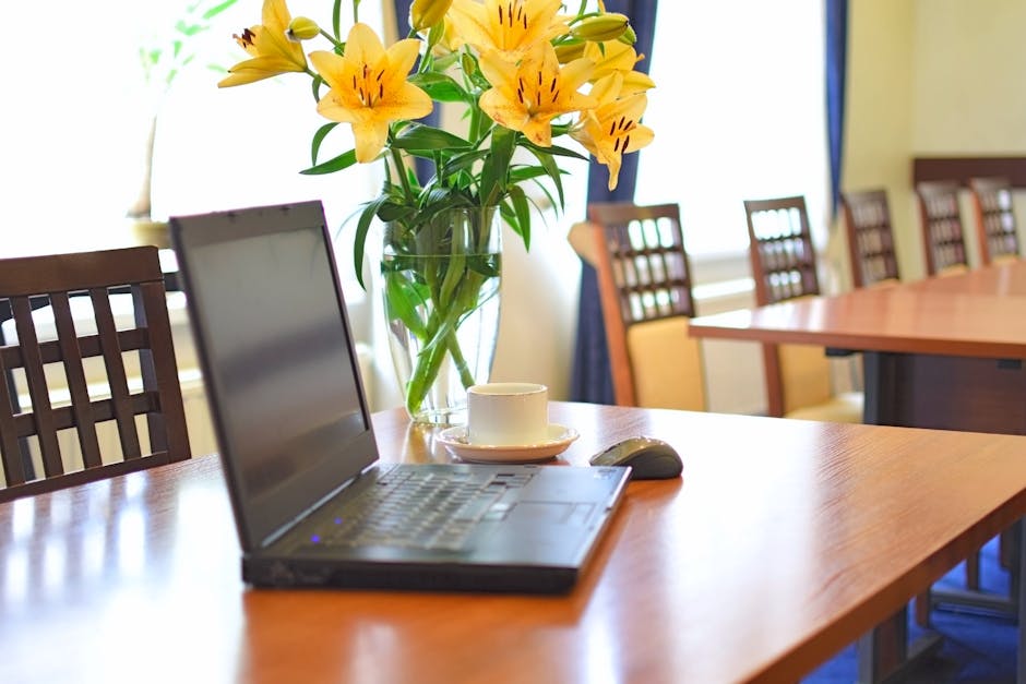 A clear glass vase filled with a floral bouquet of yellow lilies with orange streaks and closed buds, set on a polished wooden conference table. Next to the vase is a white ceramic coffee cup on a mat