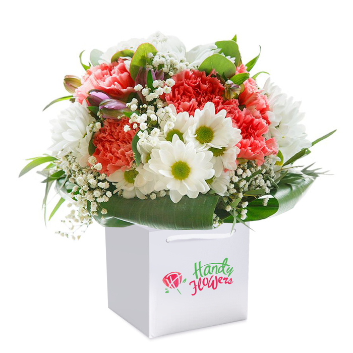 This beautifully lush bouquet, standing tall inside a crisp white square gift bag, is the epitome of contemporary Addington floral design. The composition highlights generously frilled carnations in warm peach and vibrant red hues, each petal curled and soft, radiating from the centre like a gentle embrace. Alongside, gleaming white daisies enriched by vivid lime-green eyes inject a lively freshness that echoes the energy of Addington's open spaces and green walks. Wispy white sprigs of baby's breath illuminate the arrangement with a cloud-like softness, their fine petals weaving between carnations and daisies to create texture and depth. Tucked from view, a few closed flowers with a subtle purple tinge peek outward, hinting at future blooms-an understated detail adding intrigue. The whole ensemble is cocooned by deep green, shiny foliage, leaves fanned in broad arcs to cradle and support the bouquet, forming a natural, organic collar. The white gift bag's sturdy cord handles peak from the top, ready for easy gifting or local delivery. Set against a flawless white background, each element of colour and texture is given space to shine. Crafted with care near Addington's woodlands and thriving community, this bouquet promises lasting freshness and a touch of sophisticated charm. Perfect for birthdays, anniversaries, or those impromptu gestures that brighten a day, it carries the signature style of Addington's favourite local florist-thoughtful, spirited, and always in bloom.
