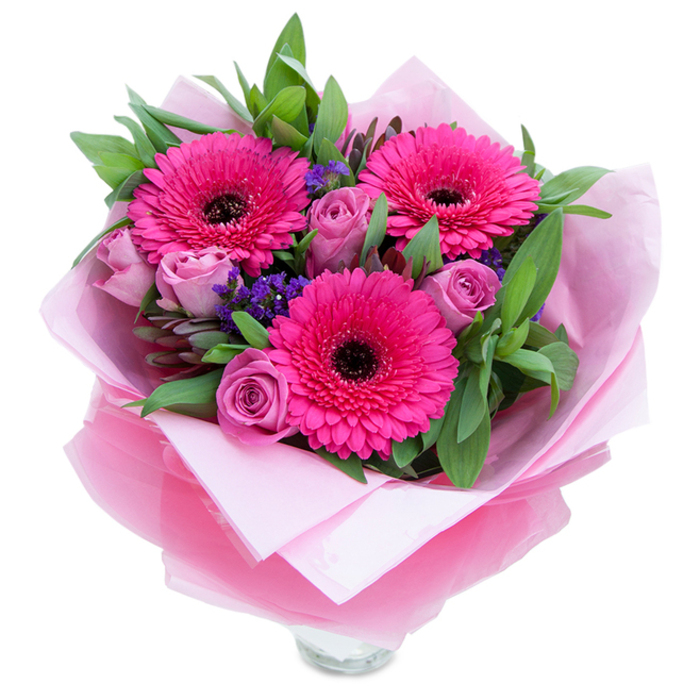 I love how this bouquet balances playful brightness with gentle grace: three intense fuchsia Gerbera daisies throw their bold, radiating petals forward, each dark-centred bloom creating a striking punctuation amid softer tones. Encircling them are roses in a muted lavender-pink or dusty rose, their petals tightly furled yet beginning to reveal the layered curves that promise fullness in the days ahead; these roses add a romantic softness and a faint, classic perfume. Little clouds of deep purple statice weave through the arrangement, their crisp, papery clusters offering texture and a complementary cool note against the warm pinks. Lush green foliage-broad, glossy leaves akin to salal or rounded eucalyptus-forms a verdant frame that enhances colour contrast and suggests a freshly gathered quality. The bouquet is wrapped as a gift: matte pale pink tissue forms a gentle cradle, topped with a translucent, crinkled fuchsia-pink cellophane that catches the light and ties the presentation together. The clear glass vase's rim is visible at the base, indicating the stems are ready for immediate display on a mantel or kitchen table. Imagined in the soft light of an Addington morning near Addington Hills or the village green, this arrangement feels intimate and carefully crafted-a perfect choice to celebrate a loved one or to bring a pocket of cheer to a local home.
